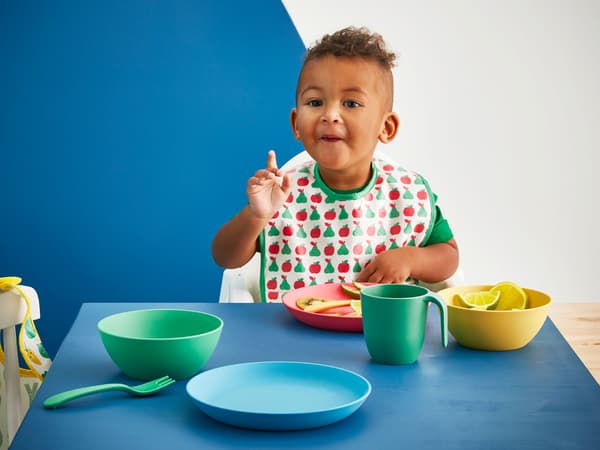 A toddler eating at a table with blue, green, yellow and red IKEA HEROISK tableware made from PLA plastic.