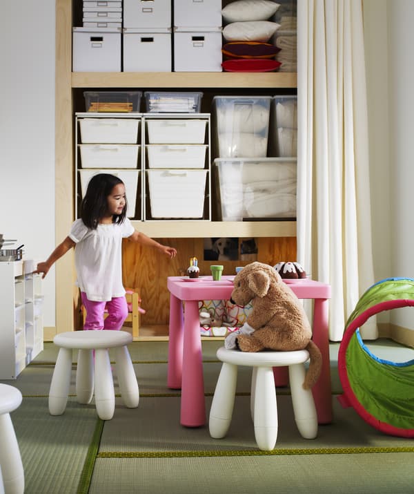 A toddler having a tea party with a soft toy, open storage in the background.