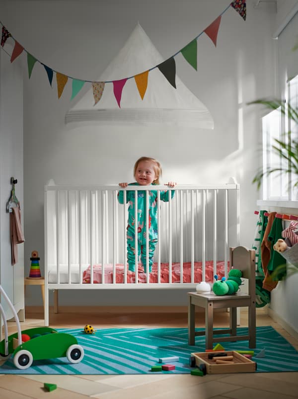 A toddler standing inside a white SMÅGÖRA cot and children’s furniture and toys in reds, greens and light blue.