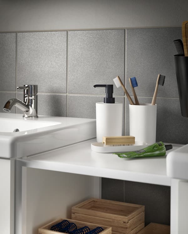A toothbrush holder with three toothbrushes, a soap dispenser and a tray in black/white stand on a shelf beside a wash-basin.