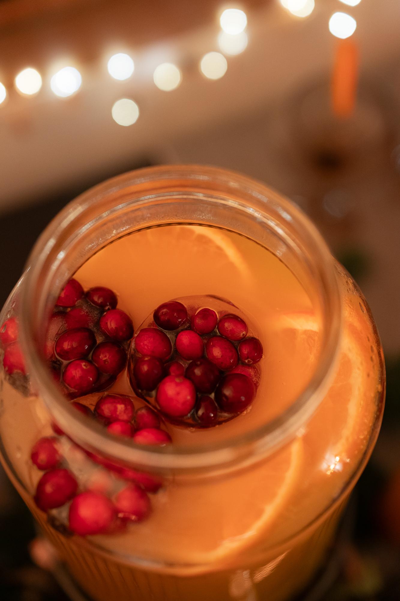A top view of a glass jar filled with a light beverage, garnished with orange slices and floating cranberries, with soft lights in the background.