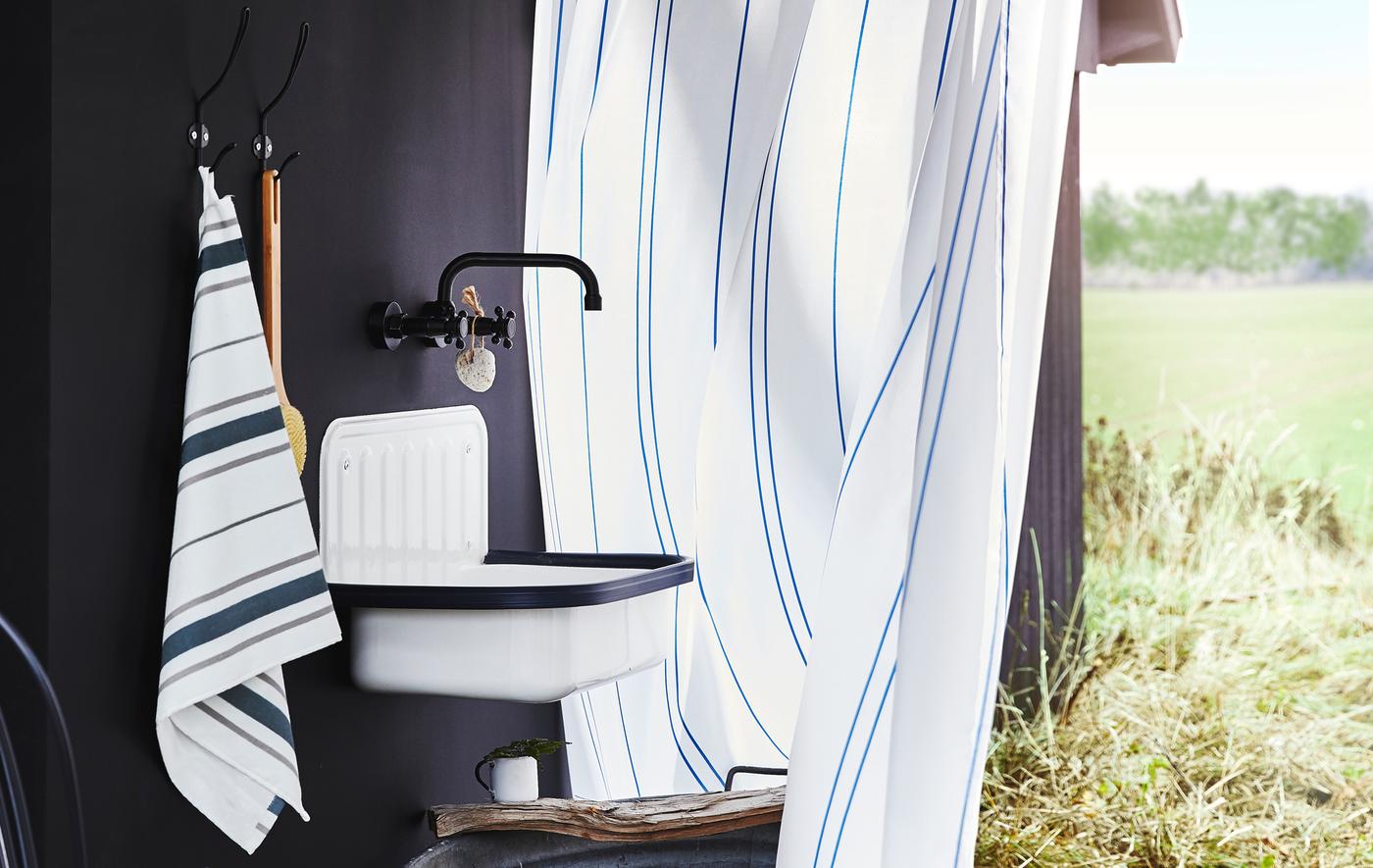 A towel, hooks, a sink and a tap on a black wall, with a field behind a striped shower curtain.