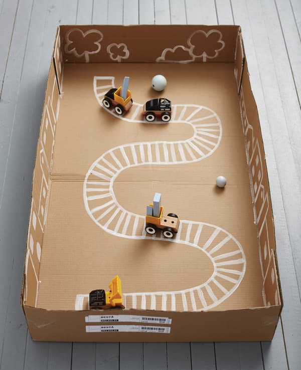 A train track is painted inside a cardboard box, with toy trains and cars.