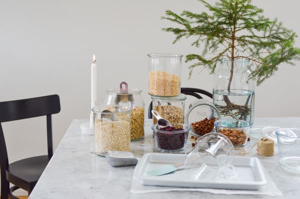 A tranquil kitchen scene with glasses filled with grains and nuts on a marble table, a burning candle, a small spruce branch in water, and a folded black chair.