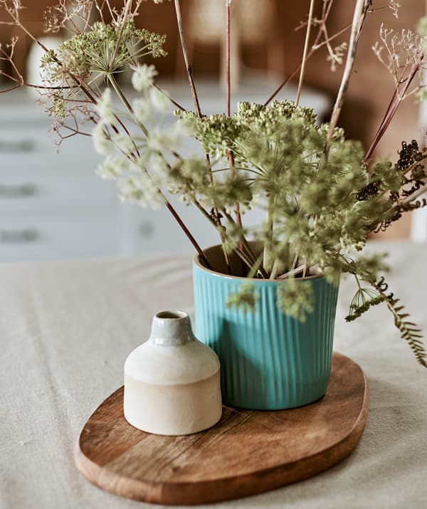 A turquoise plant pot used as a vase with a bouquet of wild leaves next to a small ceramic pot displayed on a chopping board.