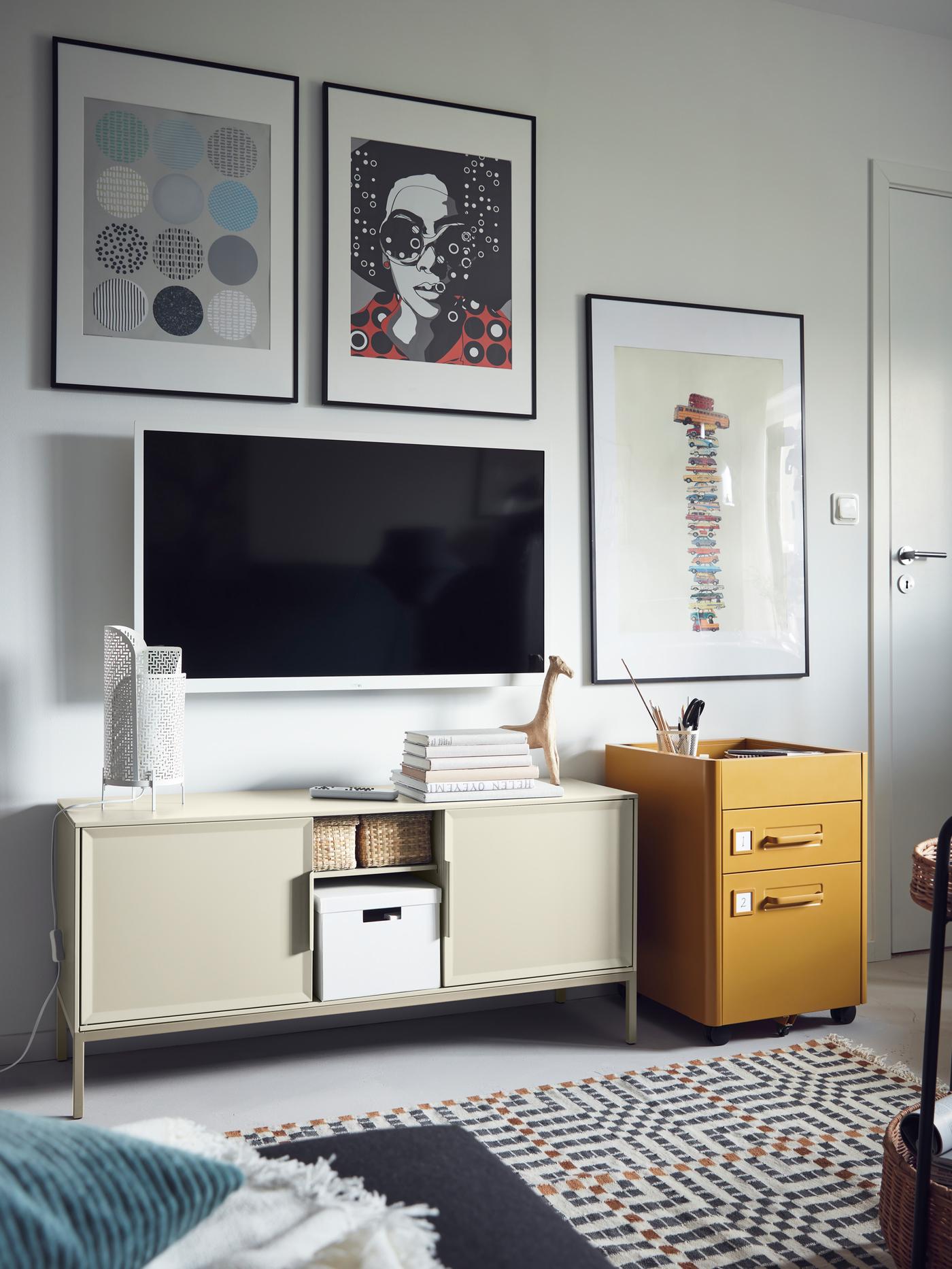 A TV and pictures are on the wall above a beige TULLSTORP metal cabinet with open storage holding baskets and a box.