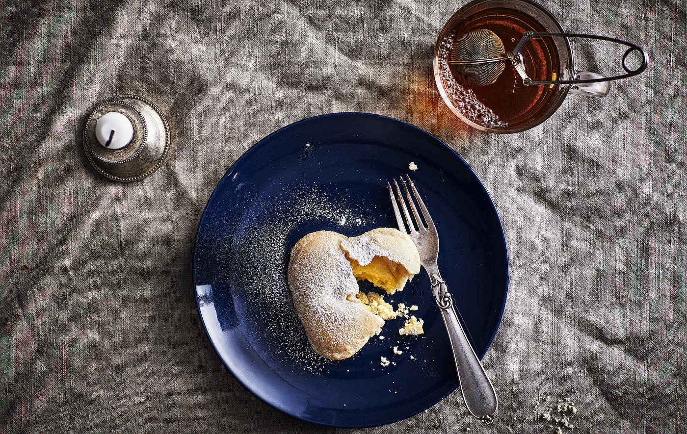 A vanilla heart biscuit and a fork on a dark blue plate, a mug with tea and a candle.