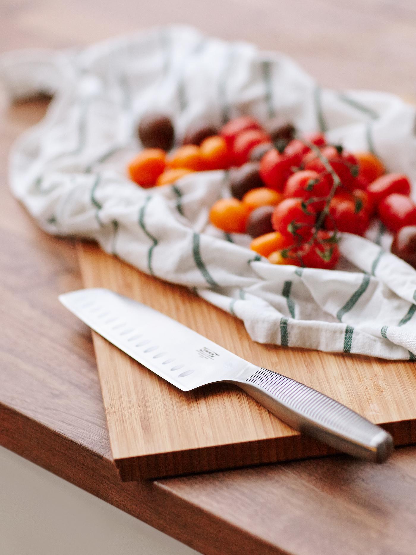 A vegetable knife in stainless steel on a wooden chopping board next to tomatoes on top of a tea towel.