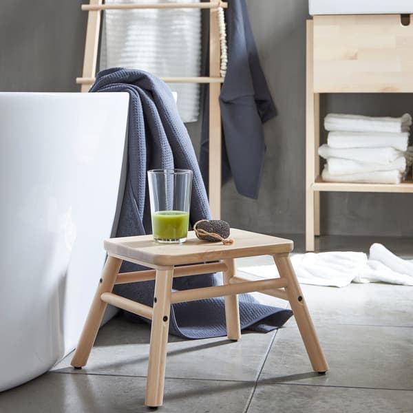 A VILTO step stool in birch wood placed beside a bathtub with a glass of green smoothie on it in a minimalistic bathroom.