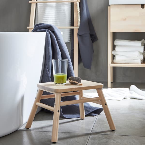 A VILTO step stool in birch wood placed beside a bathtub with a glass of green smoothie on it in a minimalistic bathroom.