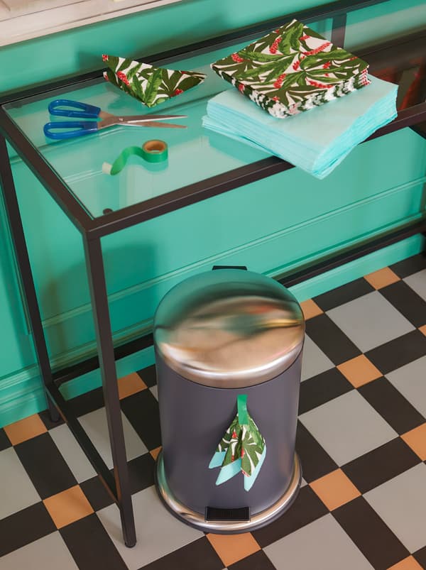 A VITTSJÖ laptop table placed along a wall, piles of FANTASTISK paper napkins on it and a MJÖSA pedal bin below.