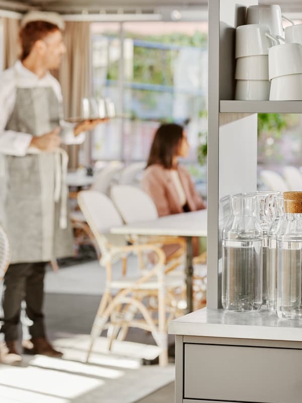 A waiter stands by a table with a woman sitting in a sunny restaurant with a storage cabinet with carafes and mugs in it.