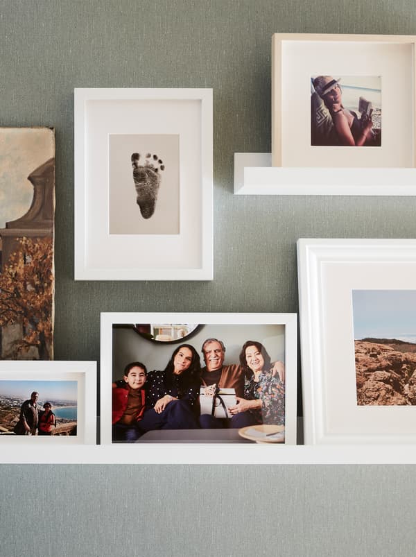 A wall with two white picture ledges and several white RIBBA frames with a baby foot print and family and travel photos.