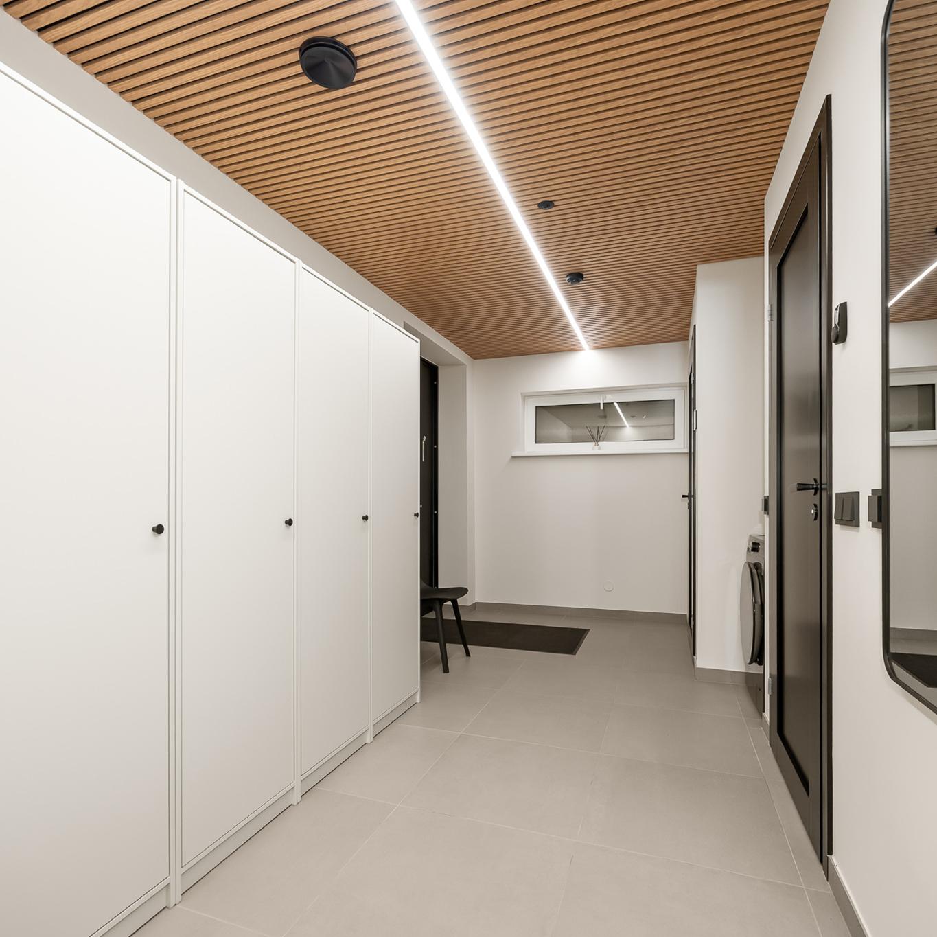 A wardrobe and changing area with light wardrobe cabinets along one wall, a large black mirror and door on the other, and a wooden slatted ceiling with a linear light. A chair and small window are visible at the far end.