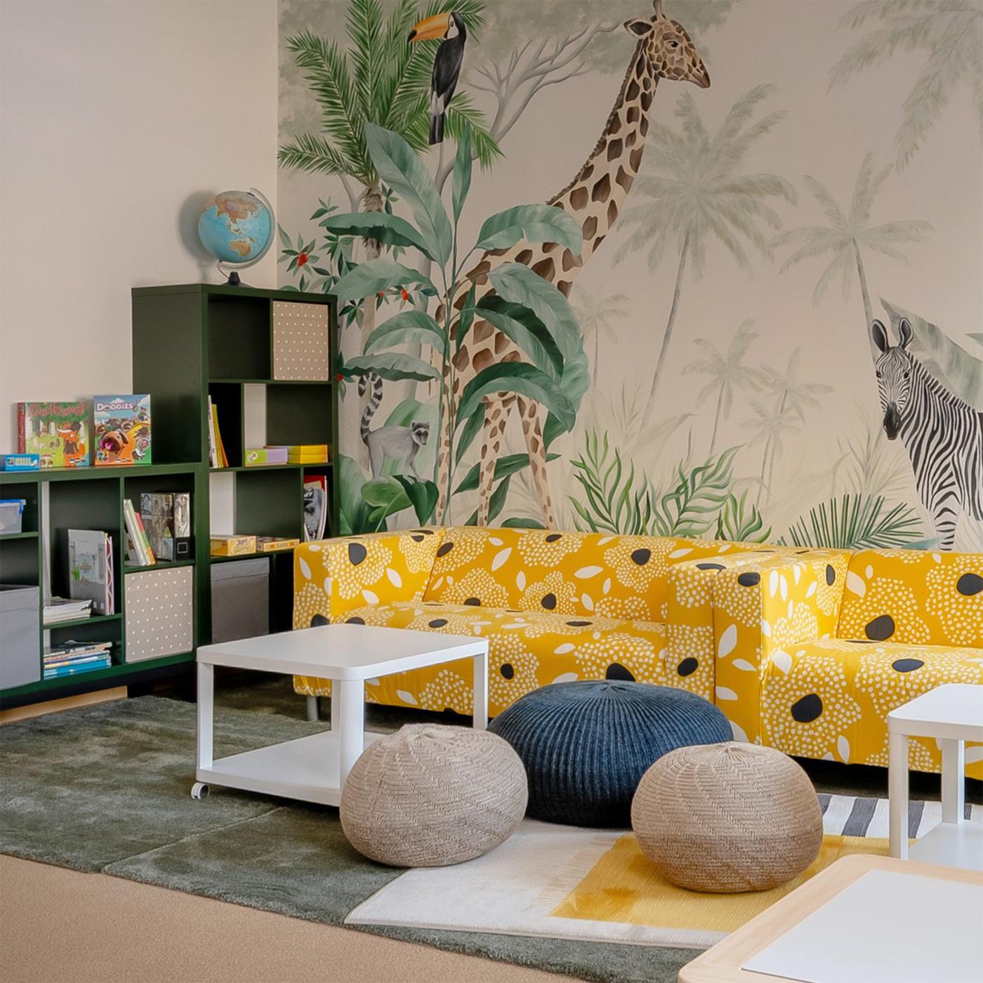 A welcoming after-school space with study tables, a lounge area, and a tropical jungle mural featuring animals such as a giraffe, zebra, and leopard. Bright yellow sofas, soft seating, and playful details create a cosy and cheerful environment for learning, relaxation, and socialising.