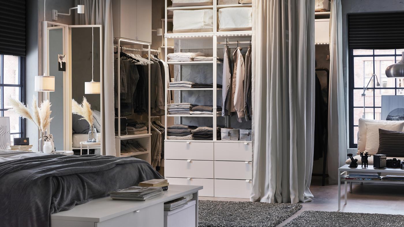 A white and grey bedroom with an open wardrobe, light grey curtains and dark grey rugs.