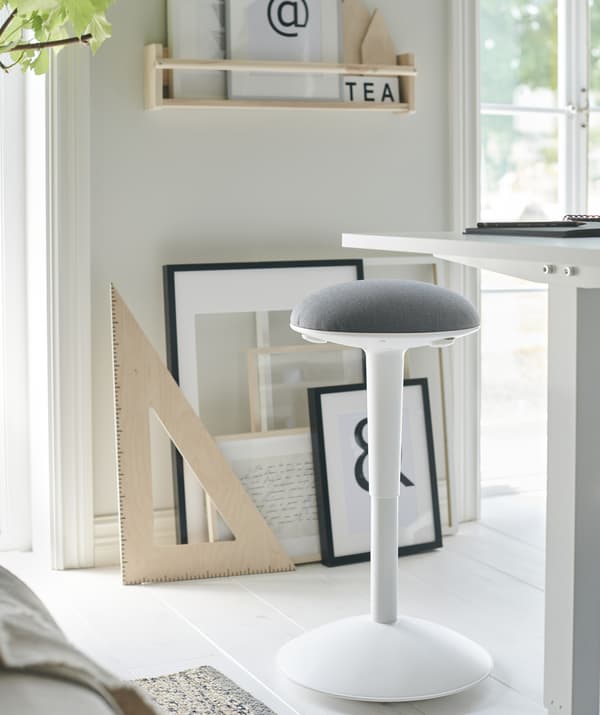 A white and grey stool next to a tall white desk, with artwork leaning against the wall behind.