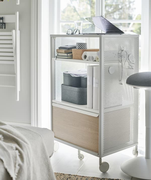 A white and light wood storage unit on wheels with a laptop on top, by a window.
