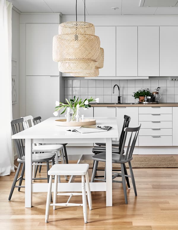 A white and neutral coloured kitchen is furnished with a large kitchen table and chairs, and decorated with a simple bouquet of flowers.