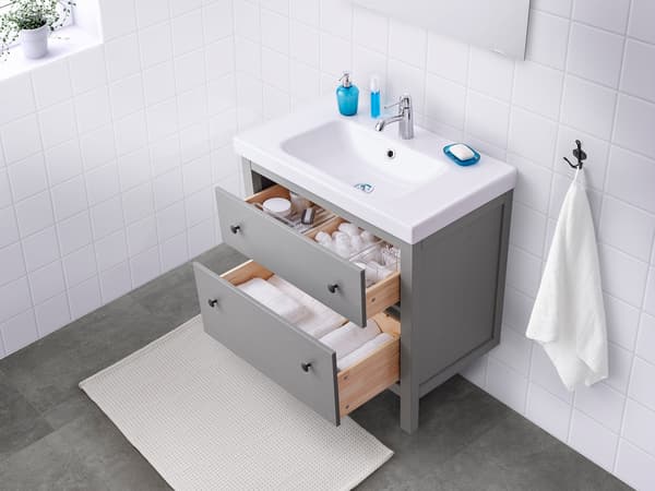A white bathroom with a gray sink cabinet with the drawers open, displaying bathroom supplies