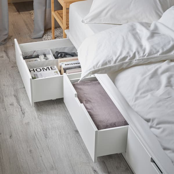 A white bed frame with two spacious, open drawers at the bottom. A pink throw, books, glasses and a watch are stored inside.