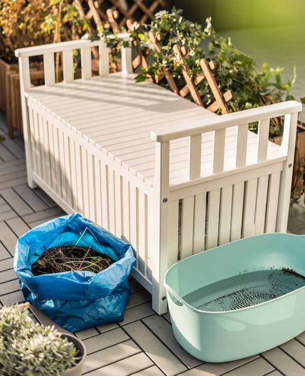 A white bench with storage, a plastic tub filled with rain water, and a blue IKEA bag filled with compost sit in the garden.