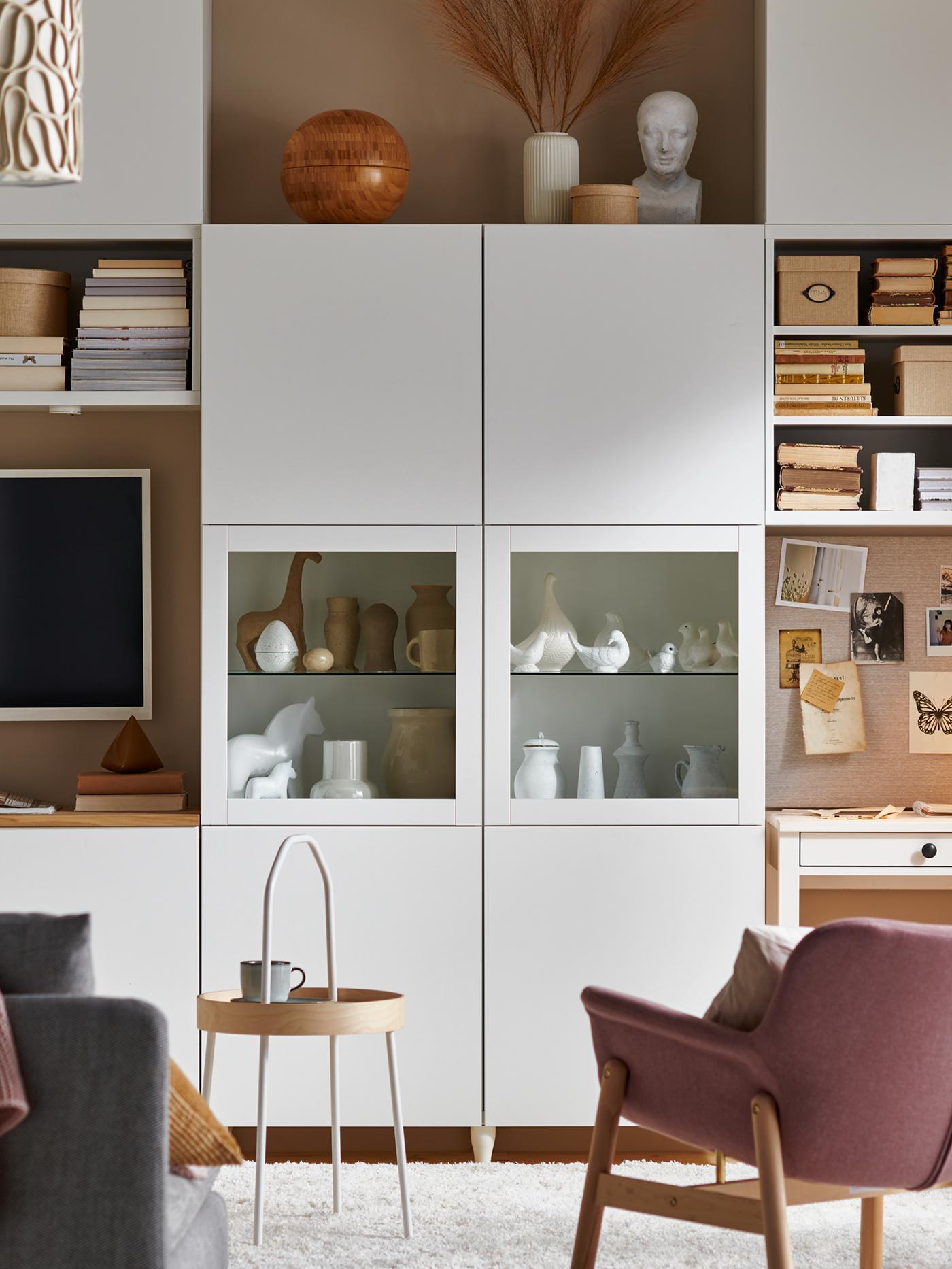 A white BESTÅ storage system showing decorative items, a TV on the wall, and a light brown-pink VEDBO armchair.