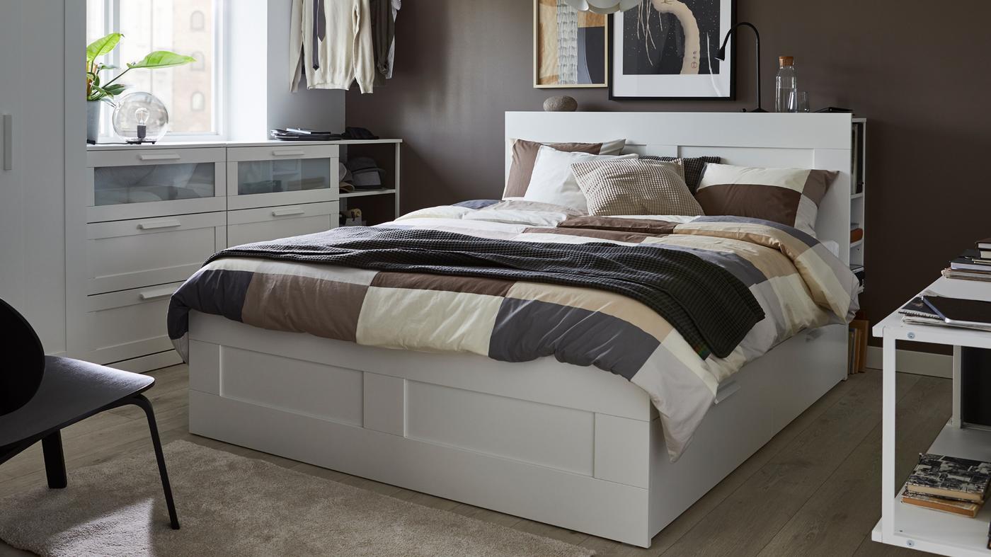 A white BRIMNES bed with storage and headboard and bed linen with a geometric pattern stands in a bedroom.
