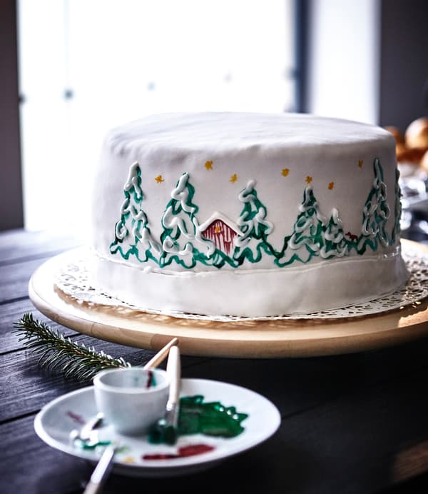 A white cake hand painted with a Christmas scene sits on an IKEA SNUDDA lazy Susan.