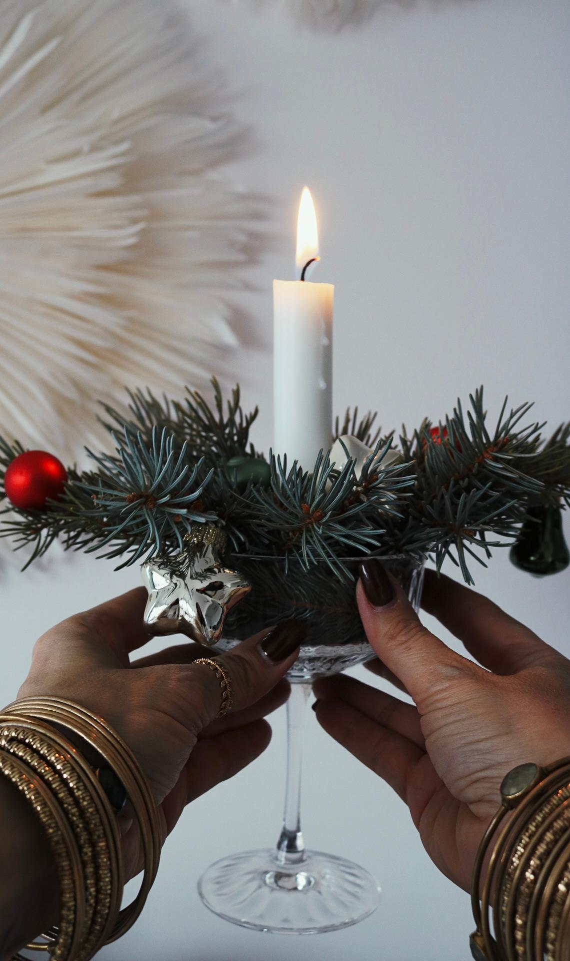 A white candle in a glass holder is adorned with pine branches and red baubles. Hands with bangles hold it, conveying a festive, elegant atmosphere.