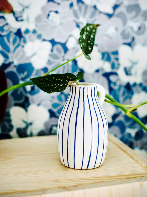 A white ceramic vase with irregular blue stripes holding a plant in front of a blue and white floral wallpaper print.