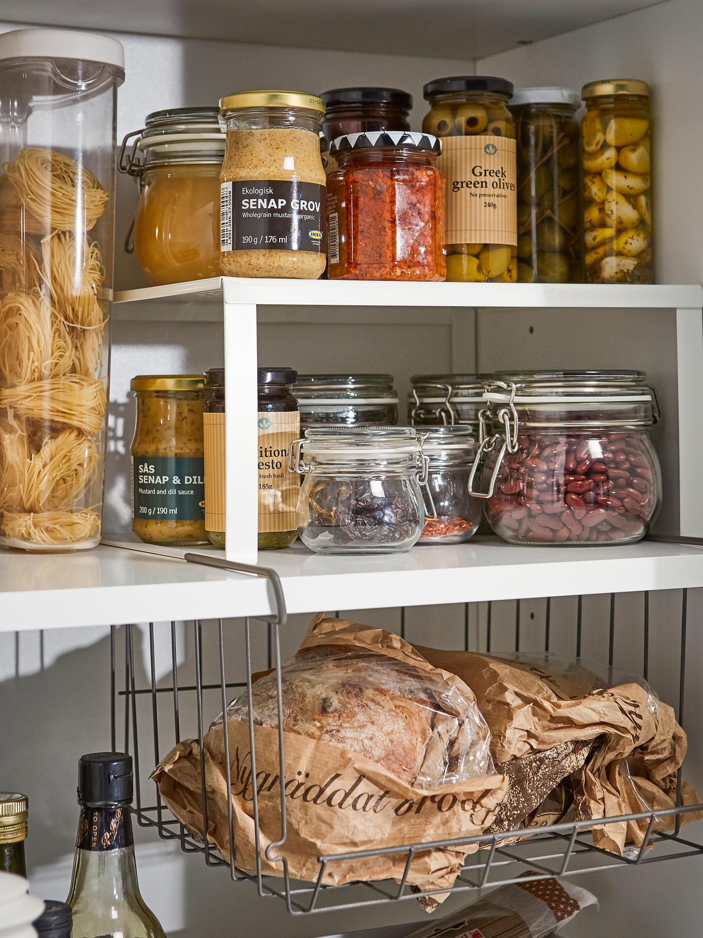 A white coated steel shelf insert and metal clip-on basket inside a pantry shelving space for jams, jars, and bottles.