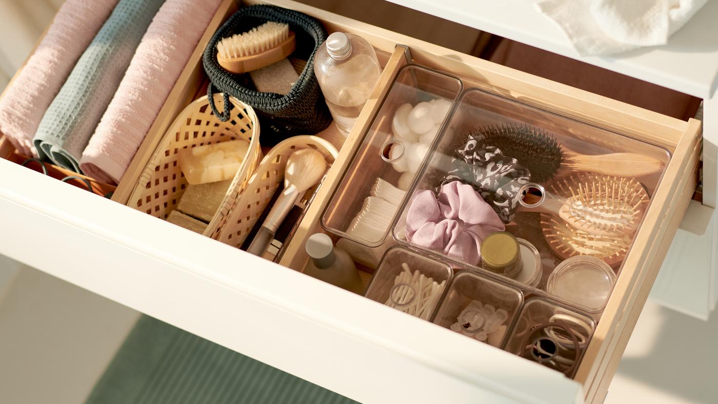 A white drawer, opened and containing transparent drawer inserts full of diverse items along with small baskets and towels.