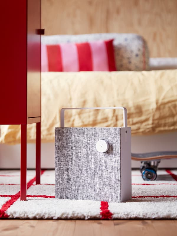 A white ENEBY Bluetooth speaker stands on a white and red rug in a modern, bright, wood-panelled bedroom.