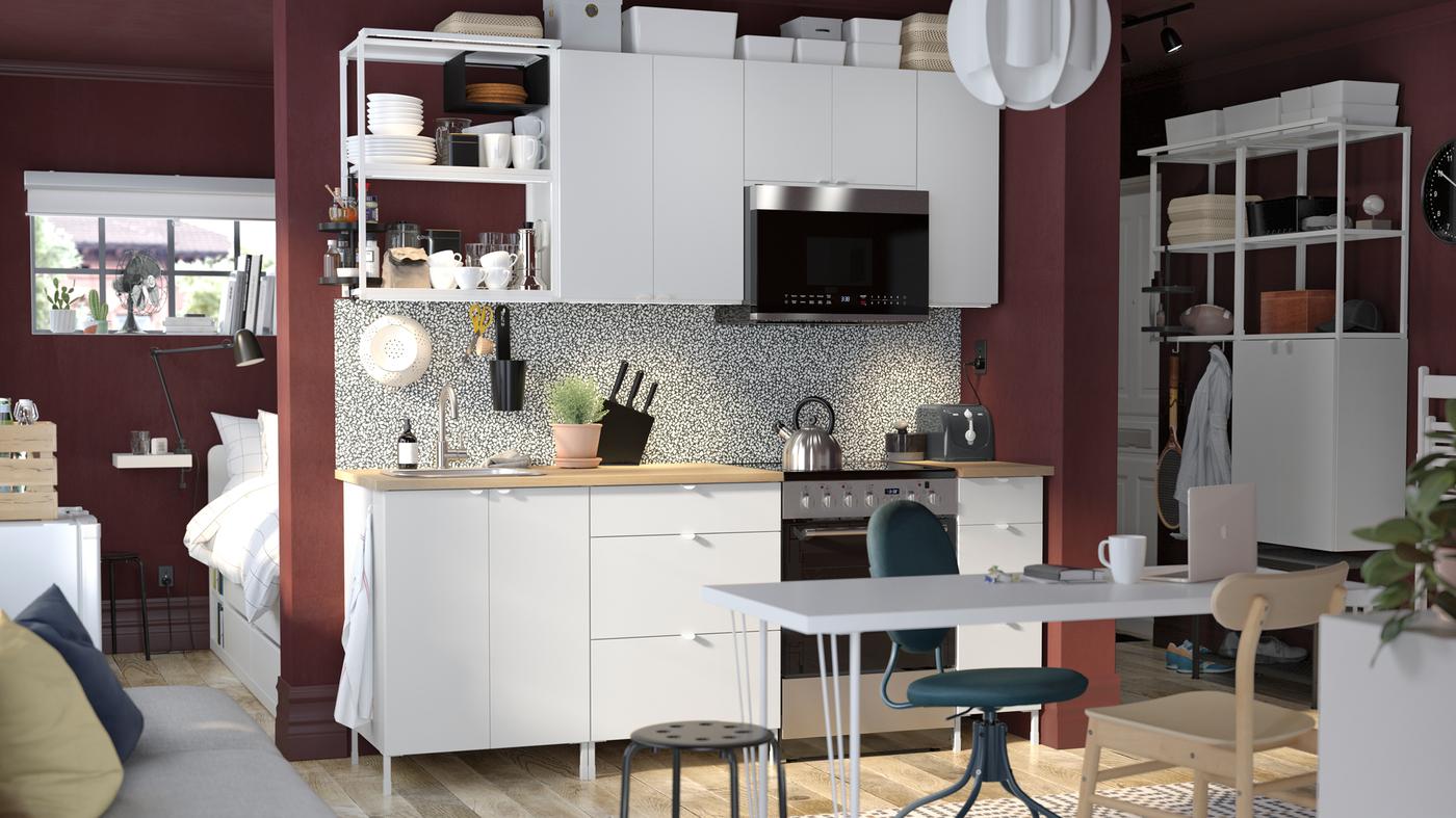 A white ENHET kitchen combination along a wall in a studio apartment with a desk in front and a bed behind the wall.