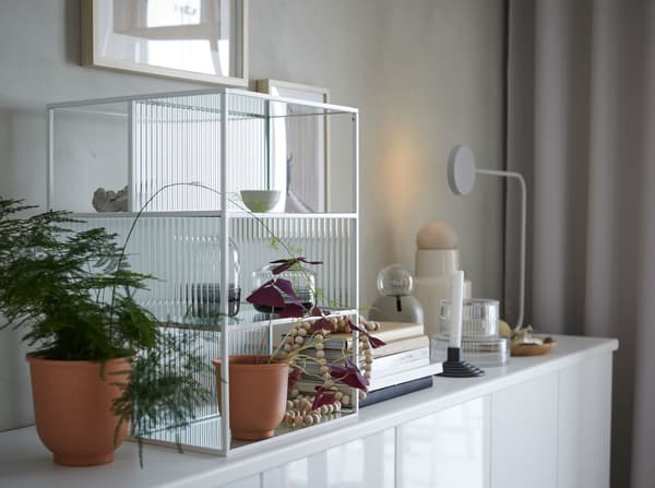 A white framed IKEA SAMMANHANG glass display box with shelves on a side cabinet with plants, pots, books and decorative items.