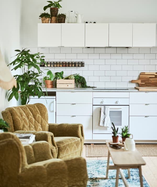 A white galley kitchen with metro tiles, two retro green armchairs and an Oriental-style rug, with a bench used as a table.