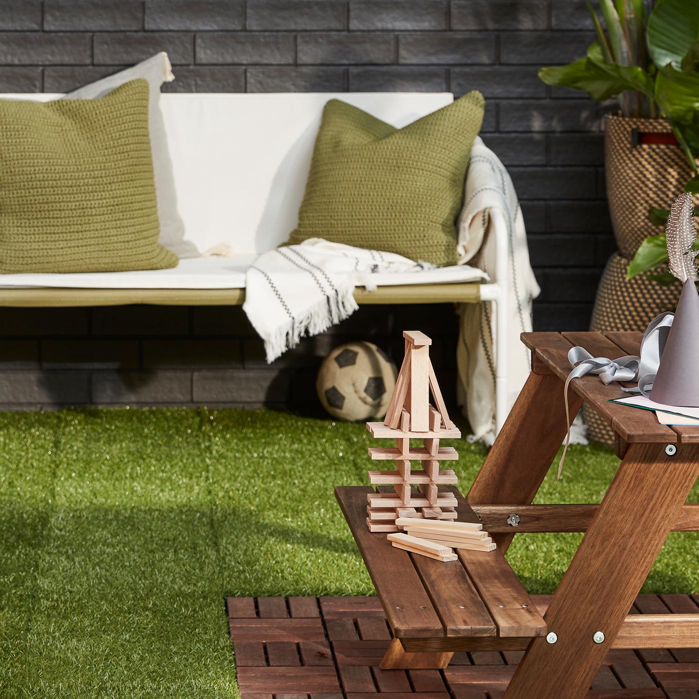 A white green/beige sofa, a potted plant and a light brown children’s picnic table on artificial grass floor decking.