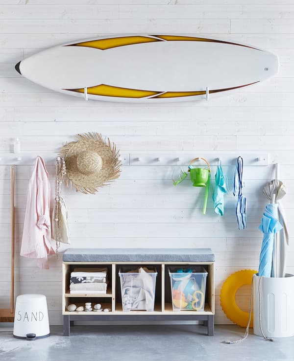 A white hall with a bench and lots of storage. A surfboard sits on a wall rack, clothes and gear hand on wall hooks.