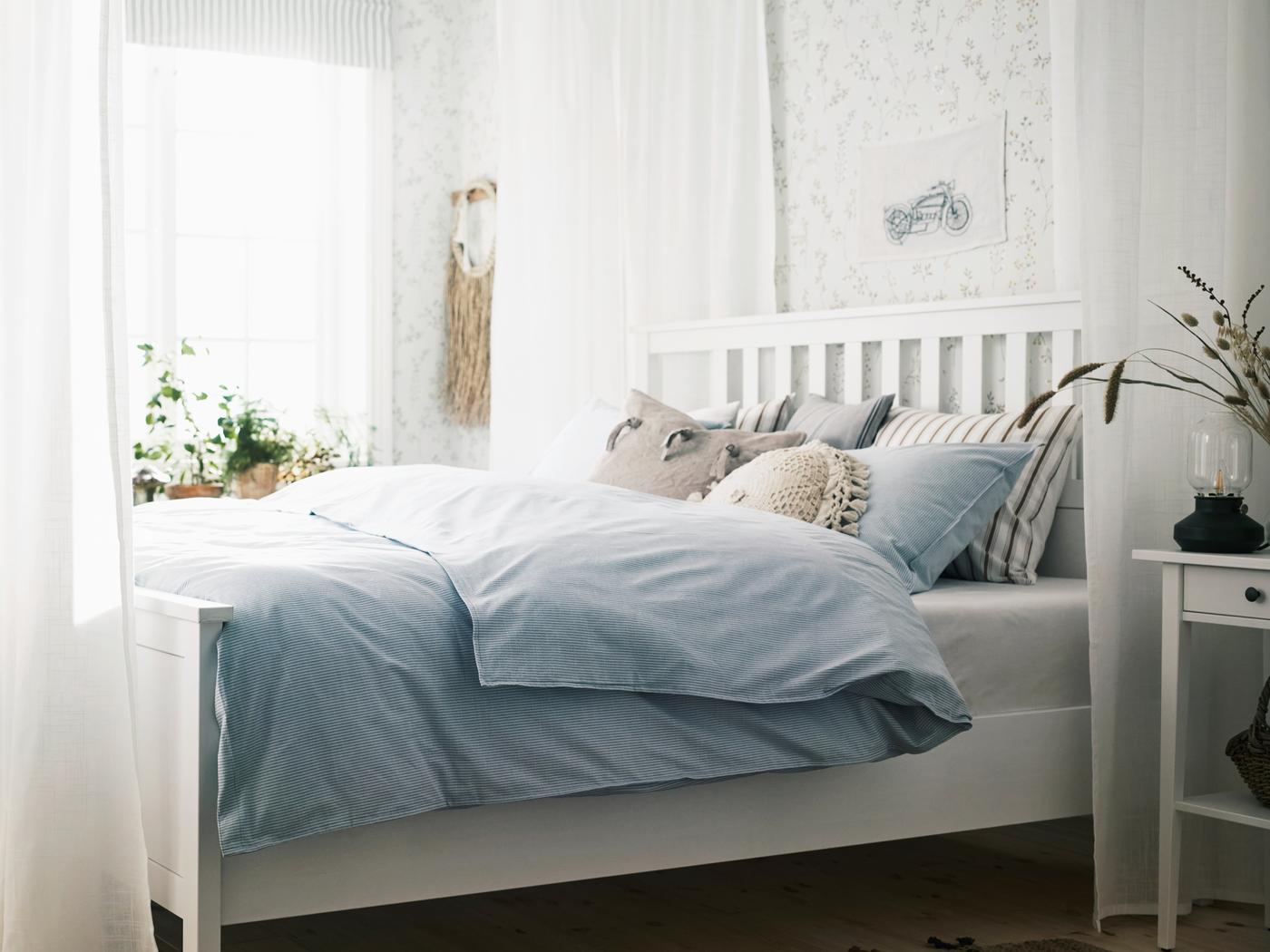 A white HEMNES bed in a bright room surrounded by sheer white SILVERLÖNN curtains, with cushions and blue bed linen on it.
