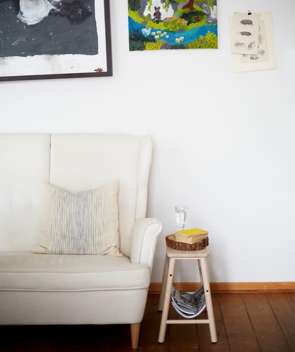 A white high-backed sofa and small wooden stool against a white wall with artwork.