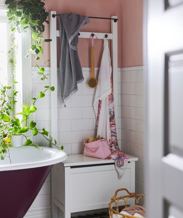 A white IKEA HEMNES storage bench with hooks and a rail next to a bath and plants.