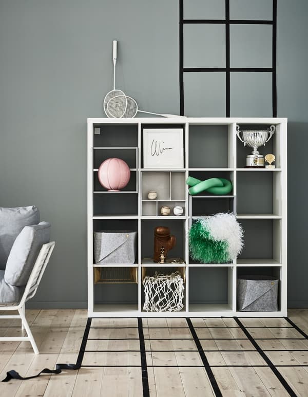 A white IKEA KALLAX shelving unit stores decorations and boxes against a grey/green wall.
