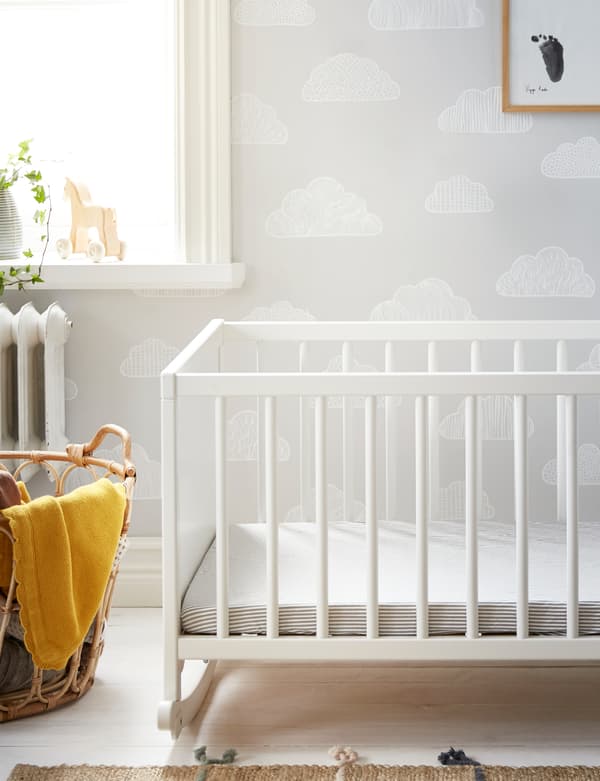 A white IKEA SOLGUL rocking crib on white wooden floorboards with SOLGUL yellow blanket and a natural textile rug.