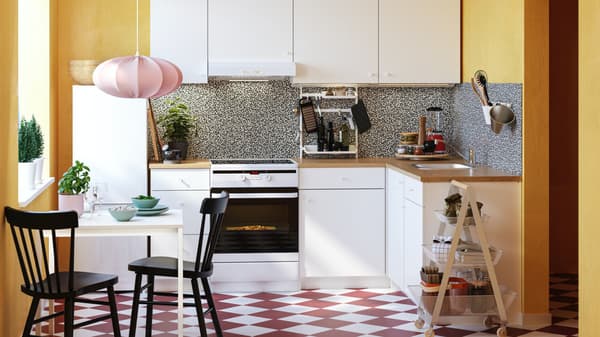 A white kitchen in a yellow room with a white and red checkered floor and black chairs at a small table with a pink hanging lamp.