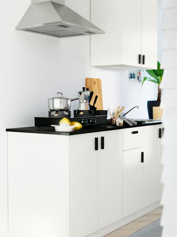 A white kitchenette with METOD cabinets, a black worktop, pots, knives, a plant and various kitchen accessories.