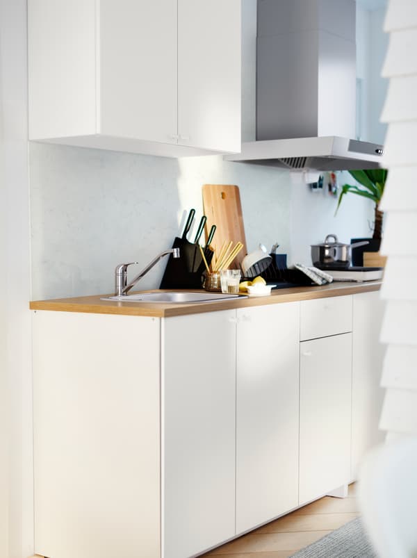 A white KNOXHULT kitchenette with a light-wood worktop, pots, knives, a plant and various kitchen accessories.