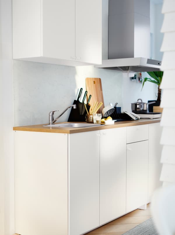 A white KNOXHULT kitchenette with a wooden worktop, potted herbs, pots, knives and various kitchen accessories.