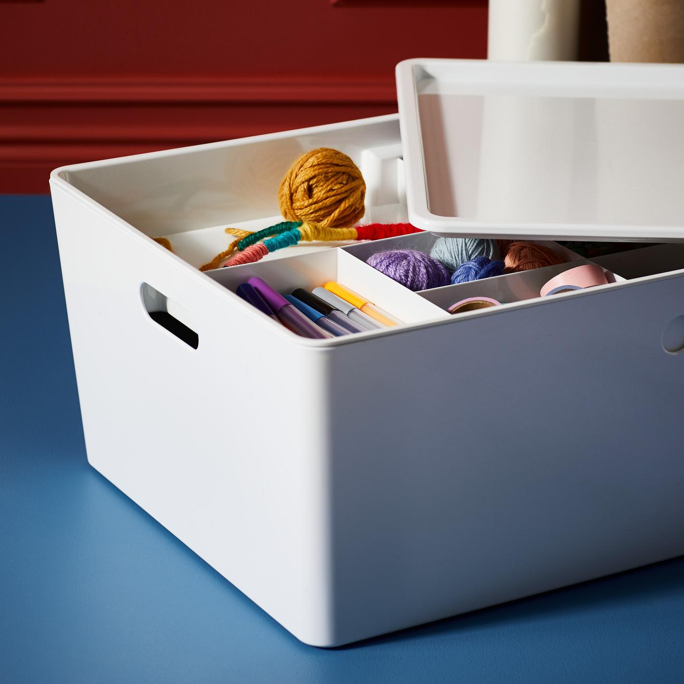 A white KUGGIS box containing a selection of coloured pens, some pink tape and several balls of brightly coloured yarn.