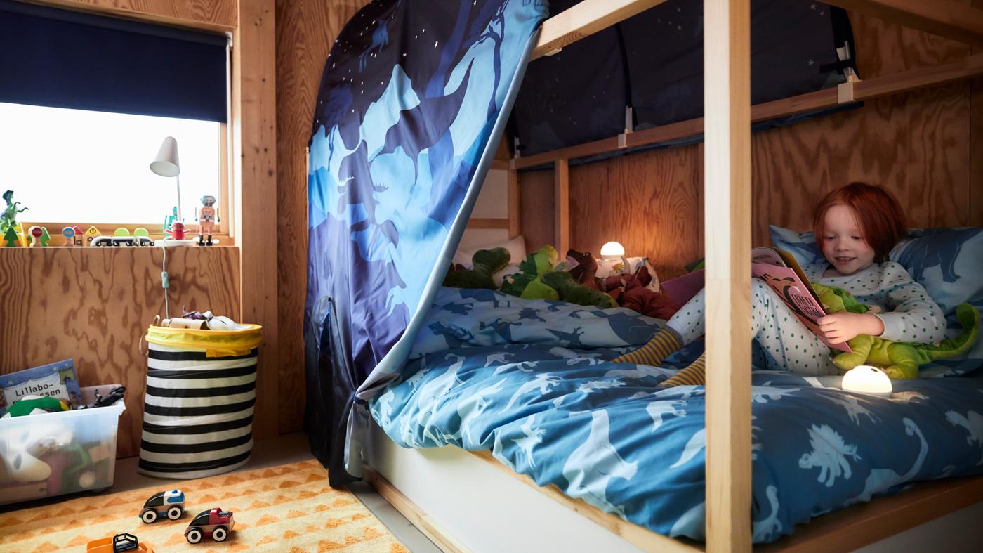 A white KURA reversible bed in a children’s room with a dinosaur themed blue tent and blue quilt cover, and a child.